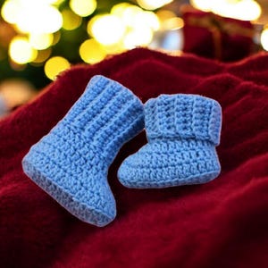 Peut inclure: Deux chaussons de bébé bleus clairs, crochetés à la main, reposent sur une surface rouge foncé texturée. Les chaussons sont faits de fil doux et ont un revers et une semelle côtelés. L'arrière-plan est flou avec des lumières de Noël.