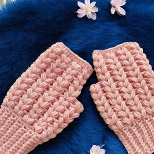Peut inclure: Une paire de mitaines roses claires avec un motif texturé et tissé. Elles sont posées sur un fond en fausse fourrure bleu vif, avec de petites fleurs blanches et roses. Accessoire douillet pour les mains.