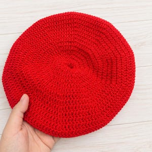 May include: A vibrant red circular beret crafted with a dense crochet stitch pattern. The hat features a uniform, textured surface created by the interlocking yarn loops, forming a consistent round shape with a small central point at the crown.