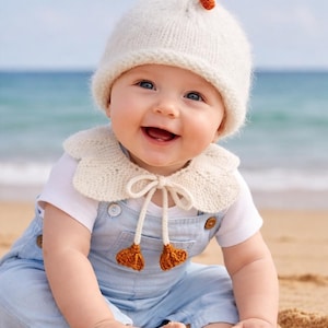 Könnte beinhalten: Ein lächelndes Baby mit einer weißen Strickmütze mit Entenmuster, einem passenden Kragen und hellblauen Latzhosen. Das Baby sitzt an einem Sandstrand mit dem Meer im Hintergrund.