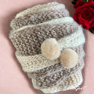 May include: A hand-knitted, beige, brown, and white striped hat with two fluffy, light brown pom-poms. The hat is made of a soft, textured yarn and is folded over. A red rose is in the background.