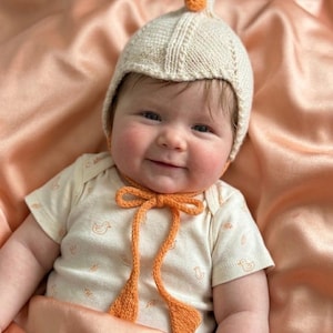 May include: A cream-colored knit bonnet featuring a small duck head on top with an orange beak, secured by long orange knitted ties. The bonnet is paired with a cream-colored short-sleeved bodysuit decorated with a repeating pattern of small orange ducks.