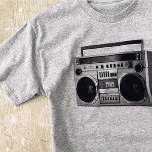 May include: A gray t-shirt with a black and white graphic of a vintage boombox.