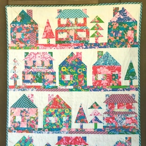 May include: A colorful quilt featuring a pattern of houses and trees. The houses are made of various fabrics with floral and geometric patterns. The trees are green and have a simple triangular shape.