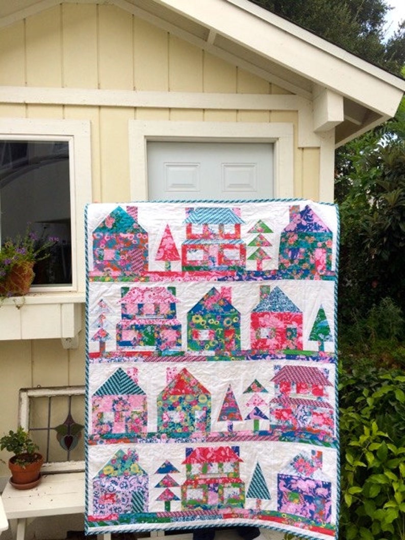 May include: A white quilt with a patchwork design featuring colorful houses and trees. The houses are made of different fabrics with floral patterns and the trees are green.