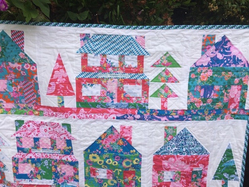 May include: A colorful quilt with a patchwork design featuring houses, trees, and the text "Hotel Fredericksburg". The quilt is made with a variety of floral fabrics in shades of pink, blue, green, and white.