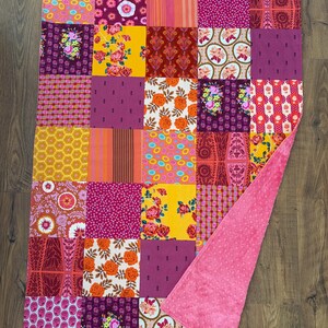 May include: A rectangular patchwork quilt featuring a variety of colorful fabric squares with floral, geometric, and striped patterns in shades of pink, orange, yellow, and purple. The reverse side is a solid, textured pink fabric with a dimpled surface.