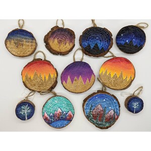 May include: Twelve round wood slices with painted scenes of mountains, sunsets, and starry nights. Each slice has a hole for hanging and is decorated with glitter and paint.