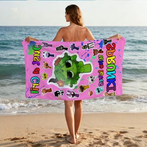 May include: A pink beach towel featuring a cartoon green character with large eyes and a flower. The towel has colorful text and various cartoon character stickers. Blue and green paint splatters are on the towel.