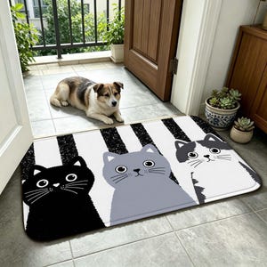 Könnte beinhalten: Eine rechteckige Fußmatte mit einem Muster aus vertikalen schwarzen und weißen Streifen. Die Matte zeigt drei illustrierte Katzengesichter: eine einfarbig schwarze Katze links, eine einfarbig graue Katze in der Mitte und eine schwarz-weiße Katze rechts.