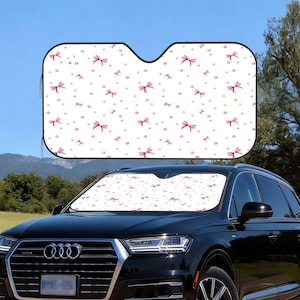 Op de afbeelding: Een witte auto zonnescherm met een patroon van roze strikken en kleine gouden stippen. Het zonnescherm is geïnstalleerd in de voorruit van een zwarte SUV en biedt bescherming tegen de zon.