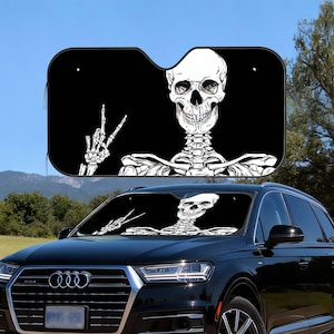 May include: Black car sunshade featuring a white skeleton graphic. The skeleton is giving a peace sign with one hand and has a smiling skull. The sunshade is designed to fit a car windshield.