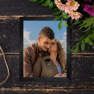 May include: A framed cross-stitch portrait of a couple embracing, with the woman kissing the man's cheek. The frame is black, and the background is a dark wood surface with flowers.