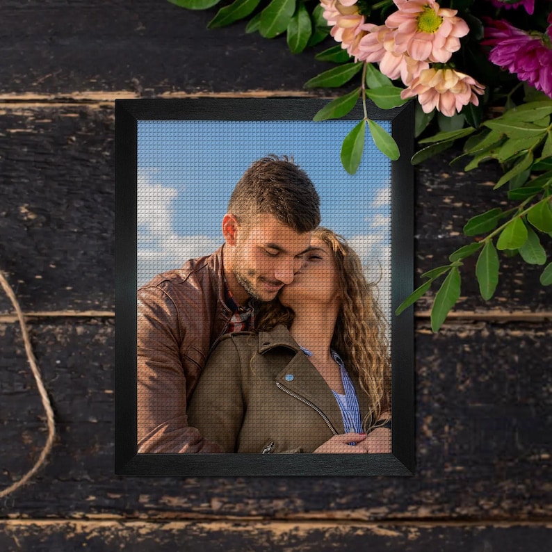May include: A framed cross-stitch portrait of a couple embracing, with the woman kissing the man's cheek. The frame is black, and the background is a dark wood surface with flowers.