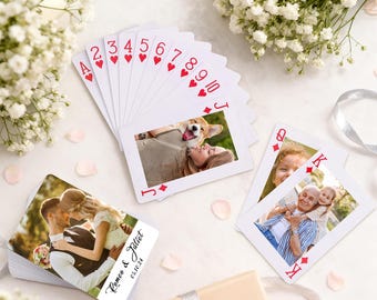May include: A deck of playing cards with custom photos, including a couple and a dog. The cards feature red hearts and the text "Romeo & Juliet" with a date. The deck is surrounded by white flowers and a gift box.