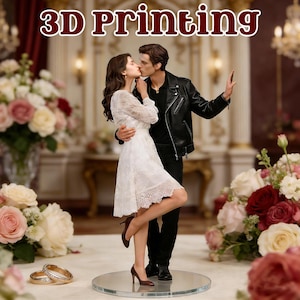 May include: A 3D-printed figurine of a kissing couple, the woman in a white dress and the man in a black leather jacket. The base is a clear, round disc. The text "3D Printing" is at the top. Two gold rings are in the foreground.