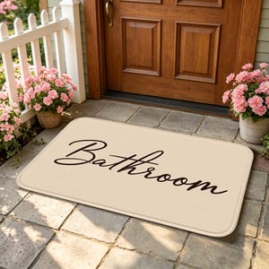 以下が含まれることがあります： 角が丸い長方形のベージュ色のフロアマットで、黒い筆記体でBathroomという文字が書かれています。マットは質感のある表面をしており、ピンクの花が飾られた木製のドアの前の石畳の上に置かれています。