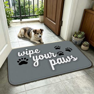 以下が含まれることがあります： 「wipe your paws」という白い筆記体のテキストと、複数の黒い肉球のグラフィックが描かれた長方形のグレーのドアマット。マットは角が丸くなっており、表面は質感のある素材で、タイル張りの床の上に置かれています。