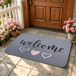 以下が含まれることがあります： 黒い筆記体でwelcomeと書かれた長方形のグレーの玄関マット。文字の下には3つのハートの輪郭が描かれており、左に白いハート、中央にピンクのハート、右に白いハートが配置されています。マットの角は丸みを帯びています。
