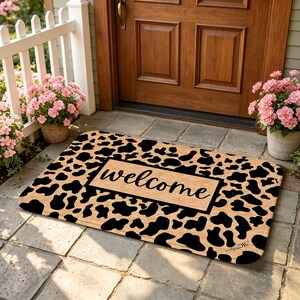 Puede incluir: Un felpudo rectangular con fondo beige y un patrón de manchas de vaca negras. En el centro, un panel rectangular beige contiene la palabra welcome escrita en cursiva negra. La alfombrilla tiene esquinas redondeadas y un borde negro.