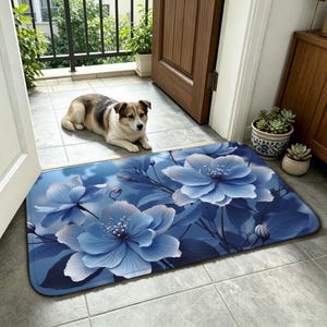 Puede incluir: Felpudo rectangular azul con grandes flores azules estilizadas y detalles blancos. La alfombra está colocada sobre un suelo embaldosado cerca de una puerta. Un perro se ve en el fondo.