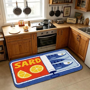 Puede incluir: Una alfombra de cocina rectangular con un diseño gráfico de una lata de sardinas abierta. El lado izquierdo es rojo con el texto amarillo SARD y dos rodajas de limón amarillas. El lado derecho es azul con tres peces estilizados.