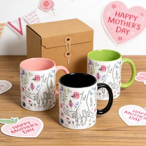 May include: Three ceramic mugs featuring a floral pattern and a line art illustration of hands holding together. One mug has a pink interior and handle, one has a black interior and handle, and one has a lime green interior and handle.