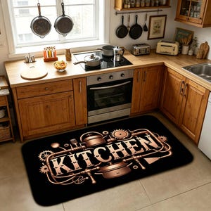 Könnte beinhalten: Eine rechteckige schwarze Bodenmatte mit einem metallisch kupferfarbenen Grafikdesign des Wortes KITCHEN, umgeben von stilisierten Rohren, Zahnrädern und Kochtöpfen. Das Design hat eine mechanische Ästhetik mit abgerundeten Ecken.