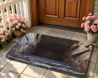 May include: Rectangular doormat with a 3D illusion of a stone staircase leading into a dark space. The mat features shades of gray, brown, and tan, creating a realistic, textured appearance. The mat is placed on a stone patio in front of a wooden door.