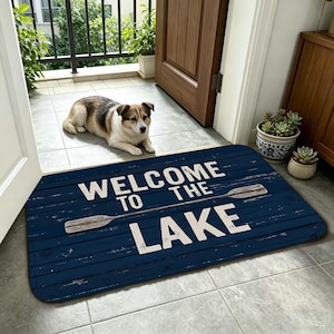 以下が含まれることがあります： 「WELCOME TO THE LAKE」の白い文字と2つのオールが描かれた長方形の青いドアマット。 ドアの前に置かれ、犬が床に横たわっています。 マットは木のような質感です。