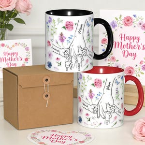 May include: Two ceramic mugs with floral designs and the words "Mom" and "Happy Mother's Day." One mug has a black handle and interior, the other a red handle and interior. The mugs feature a hand-holding design with names.