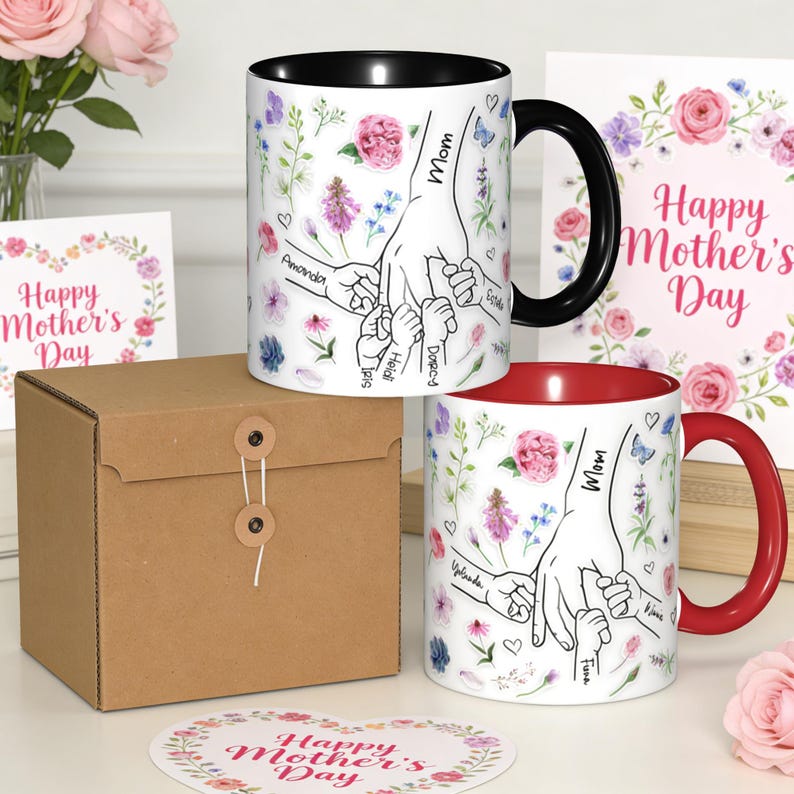 May include: Two ceramic mugs with floral designs and the words "Mom" and "Happy Mother's Day." One mug has a black handle and interior, the other a red handle and interior. The mugs feature a hand-holding design with names.