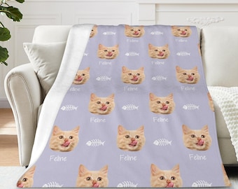 May include: A light purple throw blanket featuring a repeating pattern of orange cat faces with their tongues out, and white fish bones. The word "Feline" is printed below each cat face. The blanket is draped over a white couch.