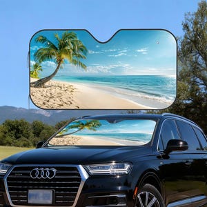 Può includere: Un'auto nera con un parasole per parabrezza con una scena di spiaggia tropicale. Il parasole mostra una spiaggia sabbiosa, palme e un oceano blu sotto un cielo luminoso. L'auto è parcheggiata all'aperto in una giornata di sole.
