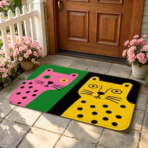 Puede incluir: Un felpudo rectangular con un diseño dividido que muestra dos caras de leopardo estilizadas. El lado izquierdo es rosa con manchas negras sobre un fondo verde, mientras que el derecho es amarillo con manchas negras sobre un fondo negro, ambos con bigotes.