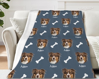 May include: A blue blanket featuring a repeating pattern of a dog's face, white dog bones, and the name "Adam" in white. The blanket is draped over a white sofa. The blanket has a white, fluffy underside.