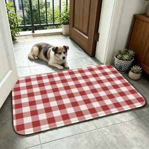 May include: A red and white checkered rectangular doormat. The mat is placed on a tiled floor near an open door. A dog is lying down on the floor near the mat. The door is made of wood and is partially open.
