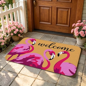 Peut inclure: Un paillasson rectangulaire à fond jaune moutarde orné de trois flamants roses et du mot welcome écrit en lettres cursives noires. Le tapis présente des coins arrondis et est posé sur une terrasse en pierre devant une porte en bois.