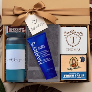 May include: A gift box with a gold bow, containing a blue tumbler, a tube of Harry's shave cream, a gray towel, a coaster with the letter "T", a bar of Dr. Squatch soap, and a Hershey's chocolate bar. The gift tag says "Gift with Love".