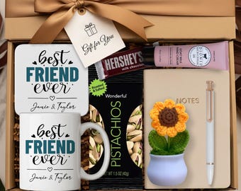 Op de afbeelding: Een cadeauverpakking met een gouden lint en een label met de tekst "Gift for You". Binnenin bevinden zich een mok en onderzetter met "Best Friend Ever", een Hershey's chocoladereep, pistachenoten, handcrème, een notitieboek, een pen en een gehaakte zonnebloem.