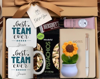 Könnte beinhalten: Geschenkbox mit goldenem Band und Anhänger mit der Aufschrift "Gift for You". Enthält eine Tasse und einen Untersetzer mit "Best Team Ever"-Text, eine Hershey's Schokoriegel, Pistazien, ein Notizbuch, Stift und eine Häkel-Sonnenblume.