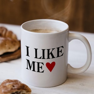 May include: White ceramic mug with a black and red design. The mug says "I Like Me" with a red heart. The mug is filled with coffee and steam is rising from the top.