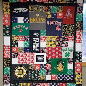May include: A patchwork quilt made of various sports team t-shirts, featuring the Boston Bruins, New England Patriots, and Boston Red Sox logos. The quilt is black with green, red, yellow, and white accents.