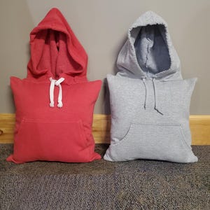 May include: Two hoodie pillows, one red and one gray, with a pocket on the front of each. The pillows are sitting on a brown carpet.