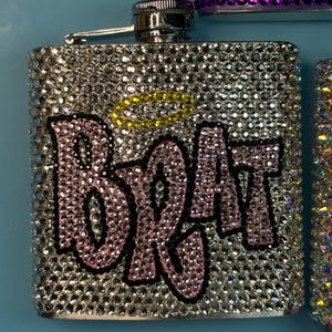 May include: A silver flask covered in sparkling rhinestones. The word "BRAT" is spelled out in pink rhinestones with a black outline. A yellow halo shape is above the word. The flask has a silver cap and hinge.