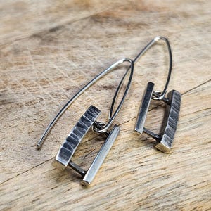 Può includere: Un paio di orecchini in argento dal design moderno. Ogni orecchino presenta due barre rettangolari collegate da un piccolo anello. Una barra ha una superficie strutturata. Gli orecchini sono esposti su una superficie di legno.