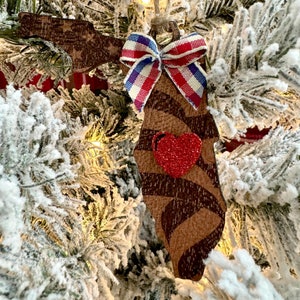 May include: A wooden ornament shaped like the state of Florida with a red glitter heart and a red, white and blue bow. The ornament is hanging on a snowy Christmas tree.