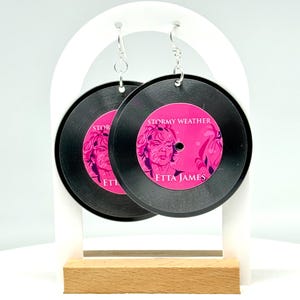 Puede incluir: Un par de pendientes de vinilo negro con una imagen rosa de Etta James y el texto "Stormy Weather" y "Etta James".