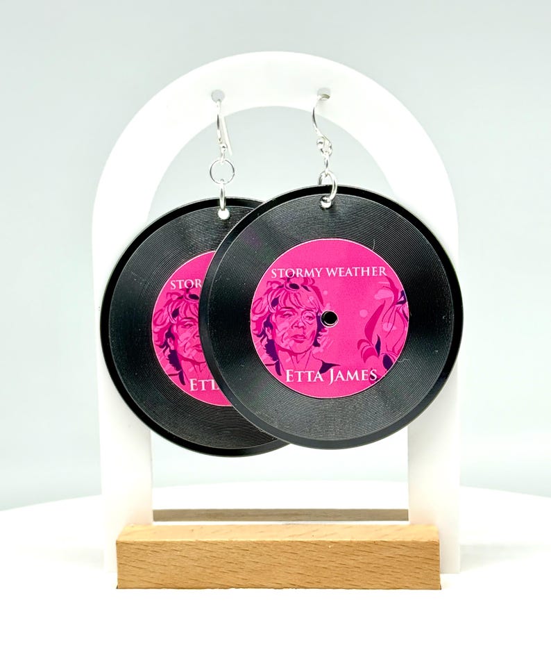 May include: A pair of black vinyl record earrings with a pink image of Etta James and the text "Stormy Weather" and "Etta James".