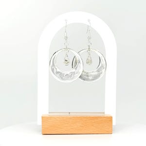 May include: Silver hoop earrings with a hammered texture and a small crystal drop dangle. The earrings are displayed on a white stand with a wooden base.